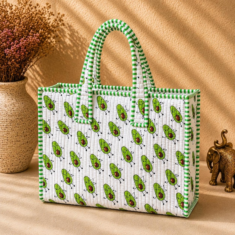 May include: A rectangular quilted tote bag featuring a repeating pattern of cartoon avocados with arms and legs on a white background. The bag has two top handles and edges trimmed with a green and white striped fabric pattern throughout.