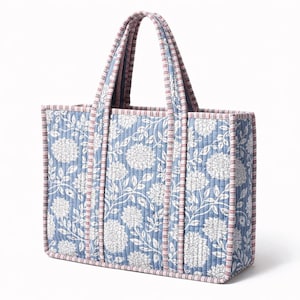 Könnte beinhalten: Eine gesteppte Tragetasche mit floralem Muster. Die Tasche hat einen hellblauen Hintergrund mit weißen Blumendesigns. Die Kanten und Griffe sind mit rosa und weiß gestreiftem Stoff verziert.