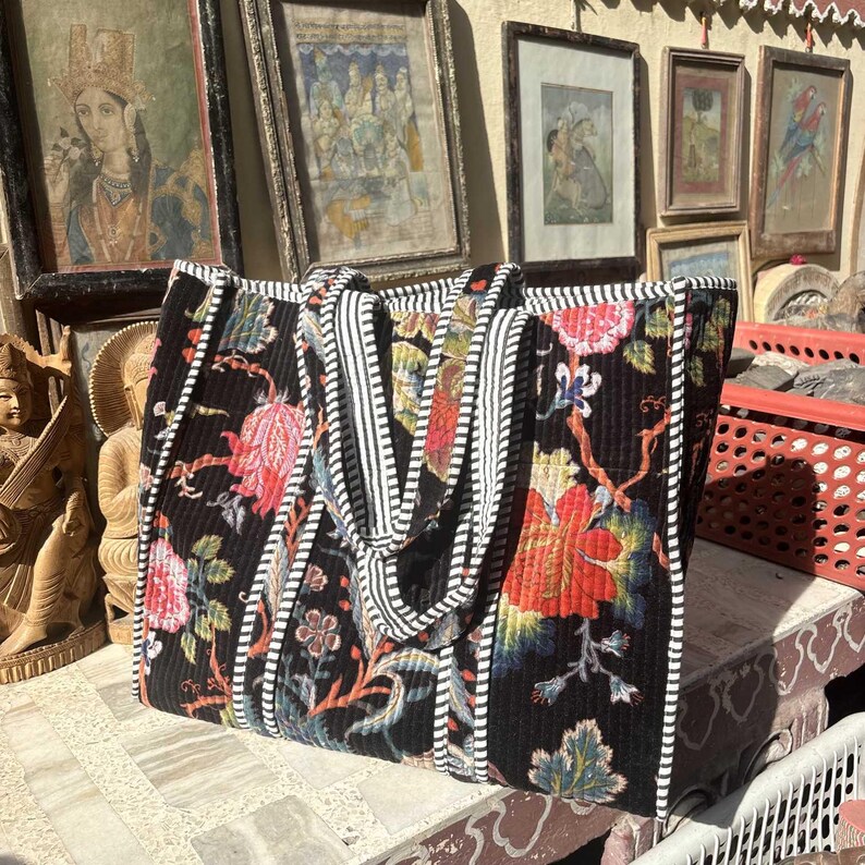 May include: A large black tote bag with a floral pattern in red, pink, and blue. The bag has white and black striped handles and trim. The bag is sitting on a stone surface.