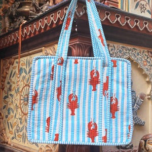 Puede incluir: Bolso tote rectangular con rayas verticales azules y blancas y un patr&oacute;n repetido de langostas rojas. El bolso tiene asas a juego a rayas azules y blancas y un aspecto acolchado, lo que sugiere una construcci&oacute;n de tela.