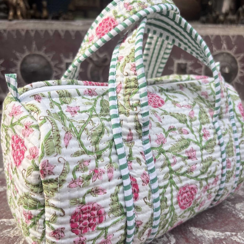 Pode incluir: Uma bolsa acolchoada de formato cil&iacute;ndrico com base branca e um padr&atilde;o floral em tons de rosa e verde. A bolsa inclui duas al&ccedil;as com um design de riscas verdes e brancas e um fecho de correr branco na parte superior.