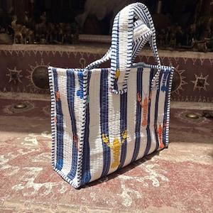 May include: A quilted tote bag featuring a vertical blue and white striped pattern with scattered orange and yellow lobster illustrations. The bag has two matching striped handles and a rectangular structure with visible stitching throughout the fabric.