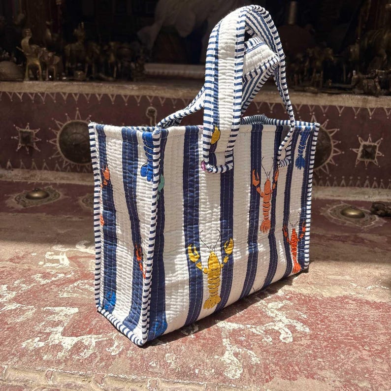 May include: A quilted tote bag featuring a vertical blue and white striped pattern with scattered orange and yellow lobster illustrations. The bag has two matching striped handles and a rectangular structure with visible stitching throughout the fabric.