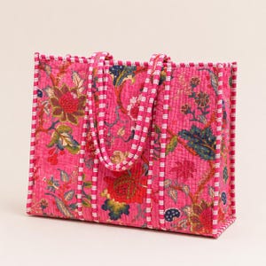 May include: A pink tote bag with a floral pattern in red, blue, and green. The bag has matching pink handles and trim with a white stripe design. The bag is rectangular and appears to be made of quilted fabric.