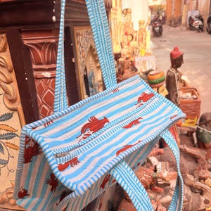 Puede incluir: Un bolso tote a rayas azules y blancas con dise&ntilde;os rojos en forma de p&aacute;jaro. El bolso tiene asas a rayas a juego y un cierre de cremallera. El bolso est&aacute; hecho de tela acolchada.