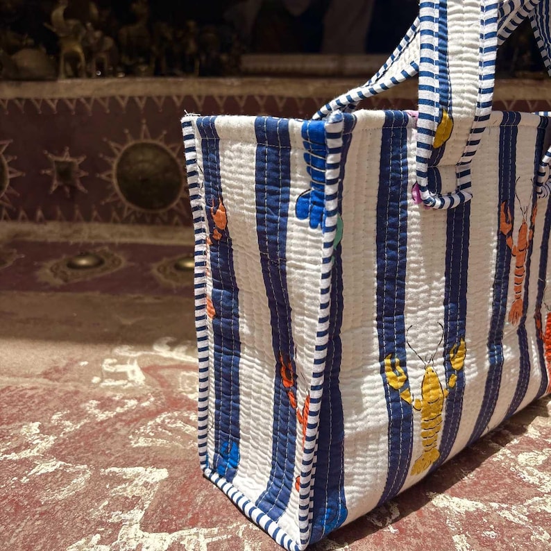 May include: A quilted tote bag featuring a pattern of vertical navy blue and white stripes. The fabric is decorated with small, colorful lobster illustrations in shades of orange and yellow. The bag includes two matching striped fabric handles.