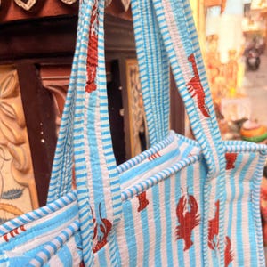 Puede incluir: Un bolso tote a rayas azules y blancas con ilustraciones de langostas rojas. El bolso tiene asas y ribetes a rayas a juego. El dise&ntilde;o es un patr&oacute;n repetitivo de langostas rojas sobre un fondo a rayas azules y blancas.