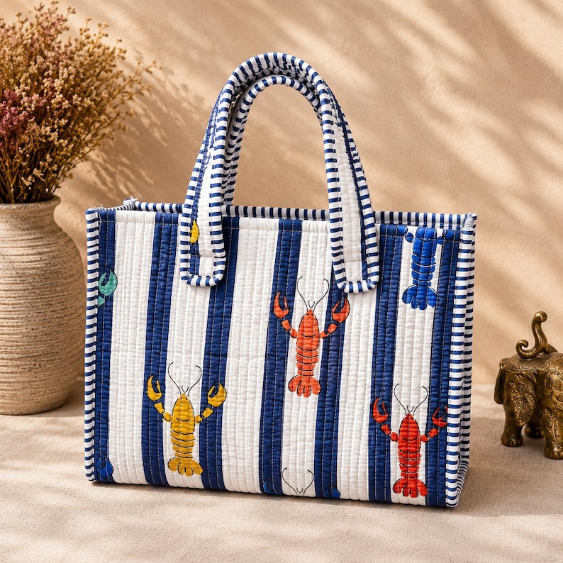 May include: A rectangular quilted tote bag featuring a pattern of vertical navy blue and white stripes. The bag is decorated with colorful lobster illustrations in red, orange, and yellow. It includes two matching striped fabric handles.