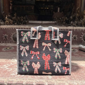 May include: A rectangular quilted tote bag in dark charcoal gray featuring a repeating pattern of red, green, and white plaid bows. The bag has two short handles with a black and white striped trim that matches the edges of the bag.