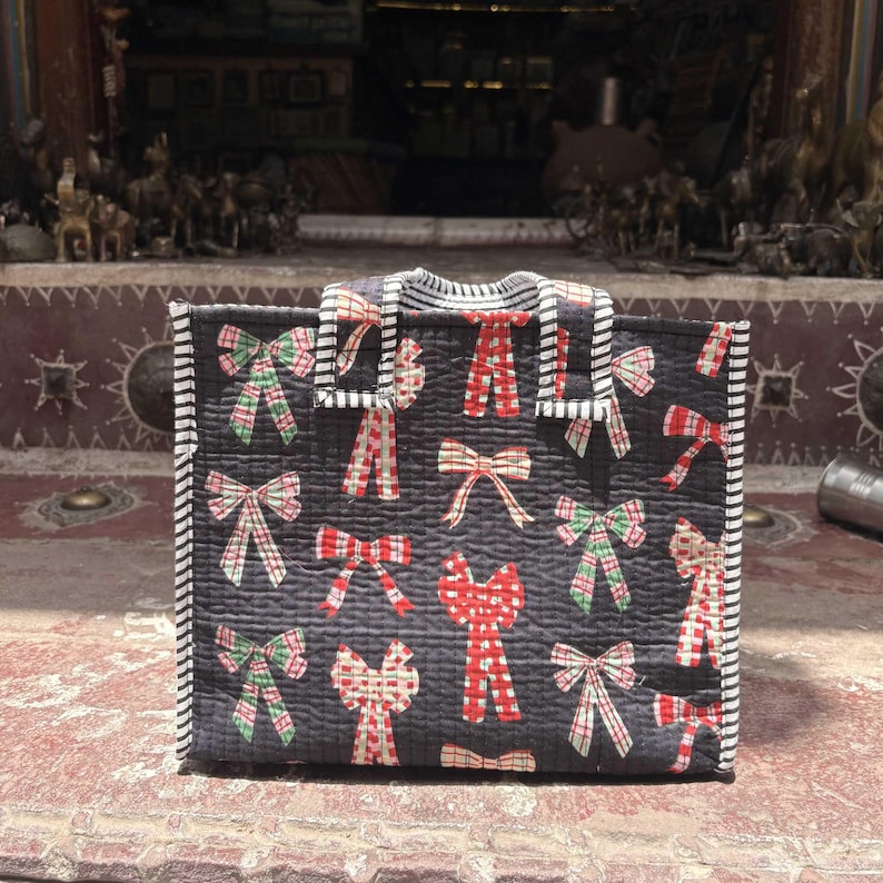 May include: A rectangular quilted tote bag in dark charcoal gray featuring a repeating pattern of red, green, and white plaid bows. The bag has two short handles with a black and white striped trim that matches the edges of the bag.