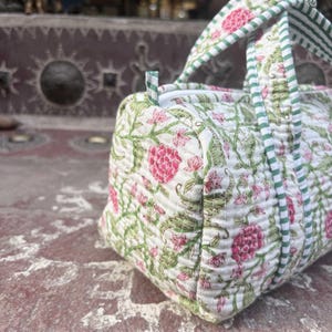 Pode incluir: Uma mala de viagem acolchoada com base branca e um padr&atilde;o floral repetitivo em tons de rosa e verde. A mala inclui al&ccedil;as de tecido &agrave;s riscas verdes e brancas e um fecho de correr superior, apresentando costuras detalhadas no tecido.
