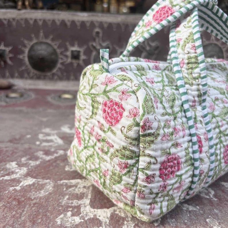 Pode incluir: Uma mala de viagem acolchoada com base branca e um padr&atilde;o floral repetitivo em tons de rosa e verde. A mala inclui al&ccedil;as de tecido &agrave;s riscas verdes e brancas e um fecho de correr superior, apresentando costuras detalhadas no tecido.