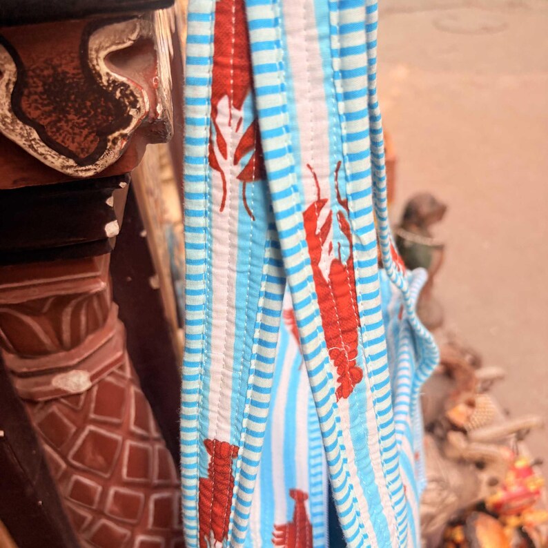 Puede incluir: Primer plano de una tela a rayas azules y blancas con dise&ntilde;os de langostas rojas. La tela est&aacute; acolchada y parece ser parte de un bolso tote o similar. El dise&ntilde;o tiene tem&aacute;tica n&aacute;utica.