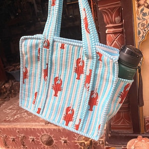 Puede incluir: Un bolso tote acolchado con un patr&oacute;n de rayas azules y blancas e ilustraciones de langostas rojas. El bolso tiene asas a juego a rayas azules y blancas y un bolsillo lateral que contiene una botella de agua verde.
