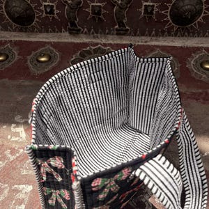 May include: A quilted tote bag featuring a black exterior with a repeating pattern of pink and green bows. The interior is lined with a black and white vertical striped fabric. The bag includes two matching quilted handles with the same striped pattern.