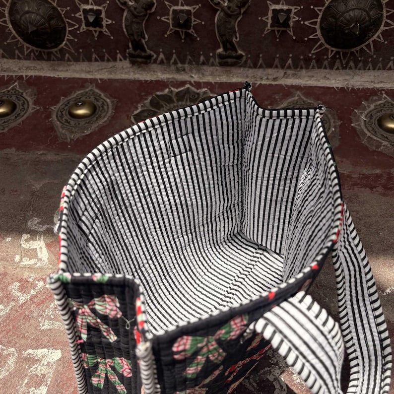 May include: A quilted tote bag featuring a black exterior with a repeating pattern of pink and green bows. The interior is lined with a black and white vertical striped fabric. The bag includes two matching quilted handles with the same striped pattern.