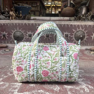 Pode incluir: Uma mala de m&atilde;o em tecido acolchoado com base branca e um padr&atilde;o floral repetitivo em tons de rosa e verde. A mala tem duas pegas superiores com um rebordo &agrave;s riscas verdes e brancas que combina com o debrum ao longo das bordas do corpo.