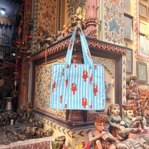 Puede incluir: Un bolso tote a rayas azules y blancas con ilustraciones de langostas rojas. El bolso tiene dos asas y se exhibe sobre un fondo de tallas y obras de arte ornamentadas. El bolso es de forma cuadrada y parece estar hecho de tela acolchada.