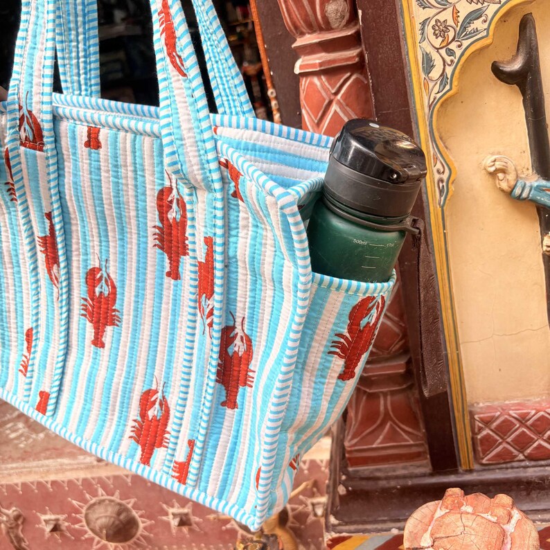 Puede incluir: Bolso tote a rayas azules y blancas con un estampado de langosta roja. Una botella de agua verde con tapa negra est&aacute; en el bolsillo lateral. El bolso tiene asas azules.