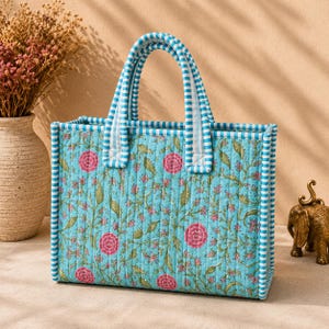 May include: A rectangular quilted tote bag featuring a light blue background with a repeating floral pattern of pink flowers and green vines. The bag has two top handles and edges trimmed in a blue and white striped fabric pattern throughout.