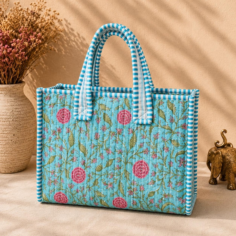 May include: A rectangular quilted tote bag featuring a light blue background with a repeating floral pattern of pink flowers and green vines. The bag has two top handles and edges trimmed in a blue and white striped fabric pattern throughout.