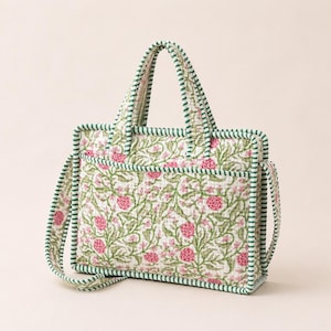 May include: A quilted tote bag featuring a floral pattern with pink flowers and green vines on a white background. The bag has two top handles and a shoulder strap, all trimmed with a green and white striped fabric along the edges and seams.