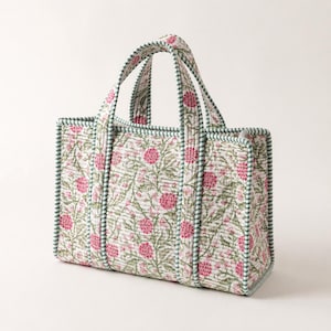 May include: A quilted tote bag featuring a floral design in pink and green on a white backdrop. The bag has two handles and is trimmed with green and white striped fabric. The rectangular bag is made of fabric and is suitable for carrying items.