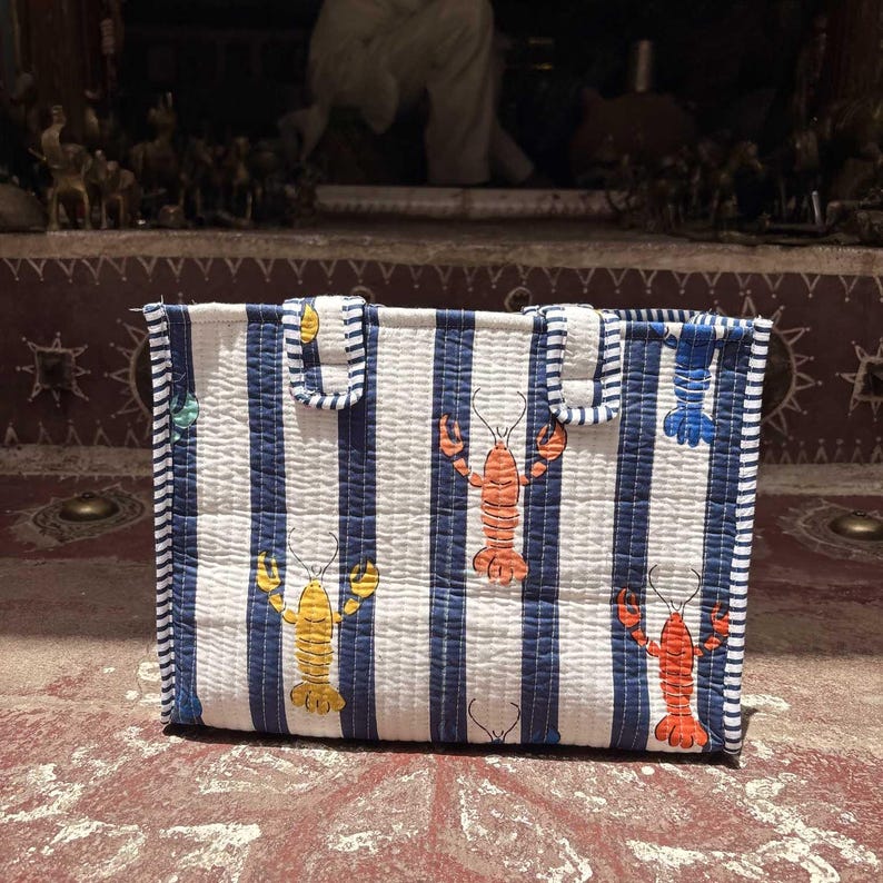 May include: A rectangular quilted tote bag featuring a pattern of vertical navy blue and white stripes. The fabric is decorated with colorful lobster illustrations in shades of orange, yellow, and red. The bag includes two matching striped fabric handles.