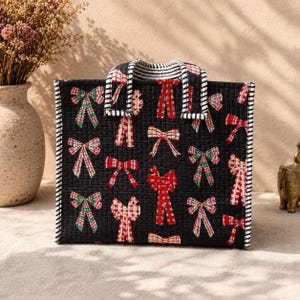 May include: A rectangular quilted tote bag with a black base featuring a repeating pattern of red, green, and white bows. The bag has two top handles with a black and white striped pattern, which also trims the vertical edges of the bag.