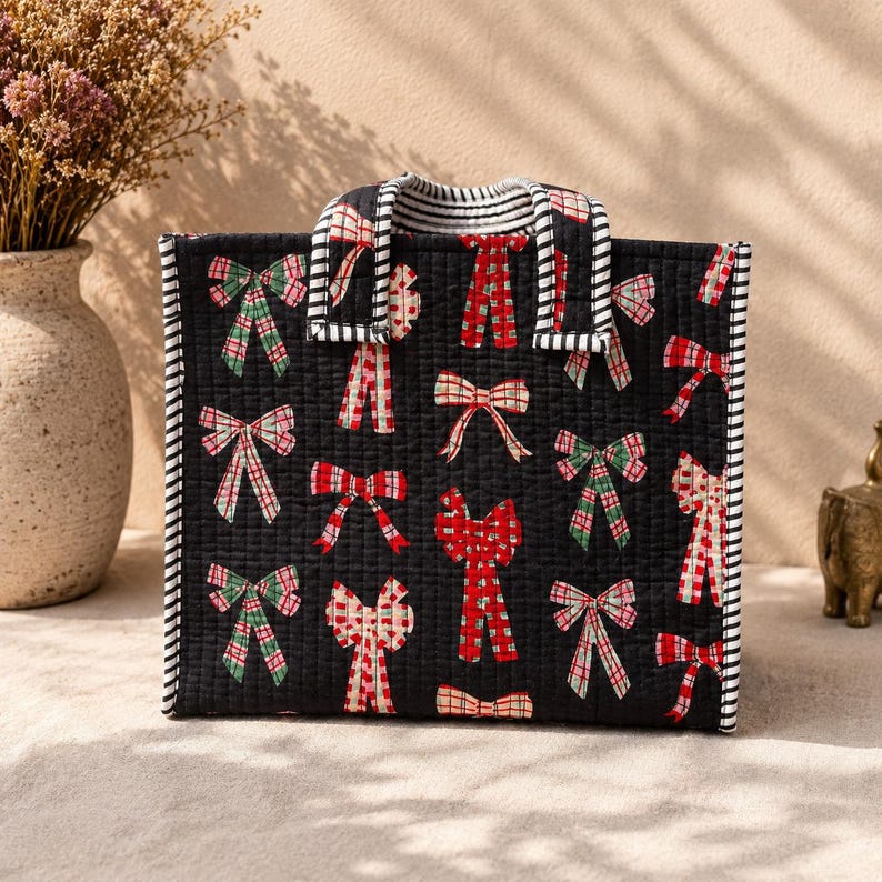 May include: A rectangular quilted tote bag with a black base featuring a repeating pattern of red, green, and white bows. The bag has two top handles with a black and white striped pattern, which also trims the vertical edges of the bag.