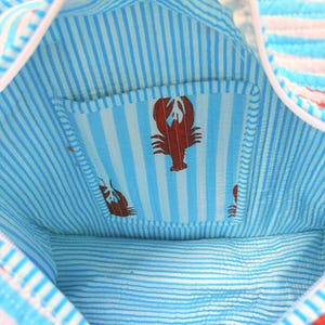 Puede incluir: Vista interior de un bolso tote a rayas azules y blancas. El bolso presenta un peque&ntilde;o bolsillo con un patr&oacute;n de rayas azul claro y blanco y un dise&ntilde;o de langosta roja. El interior del bolso est&aacute; forrado con un patr&oacute;n de rayas similar.