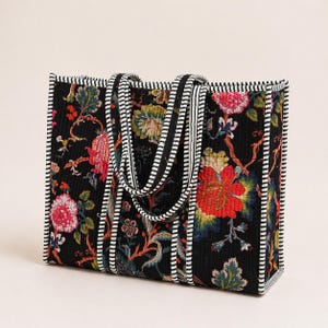 May include: A rectangular tote bag with a black background and a floral pattern in red, pink, yellow, and green. The bag has black and white striped handles and trim. The bag is quilted.