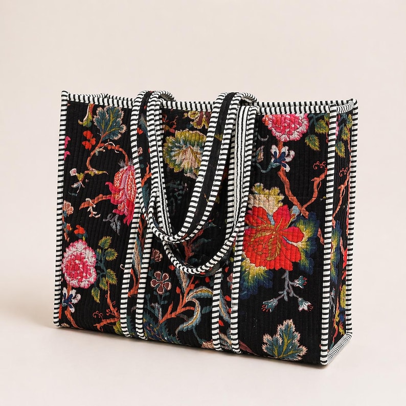 May include: A rectangular tote bag with a black background and a floral pattern in red, pink, yellow, and green. The bag has black and white striped handles and trim. The bag is quilted.