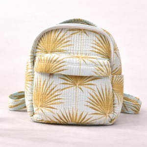 May include: A small, light blue backpack with a repeating pattern of gold palm fronds. The backpack has a front pocket, side pocket, and adjustable straps. The bag is made of a quilted fabric.