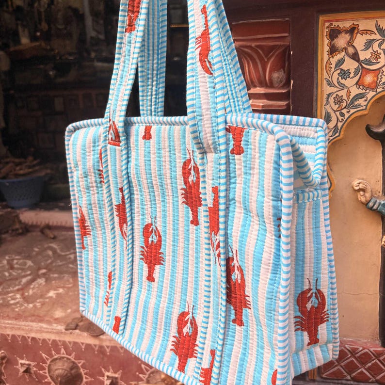 Puede incluir: Un bolso tote acolchado con un patr&oacute;n de rayas azules y blancas y un dise&ntilde;o repetido de langostas rojas. El bolso tiene dos asas resistentes y est&aacute; hecho de un material suave y acolchado. Dise&ntilde;ado para transportar objetos.
