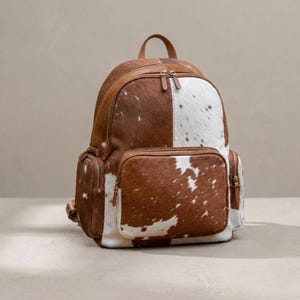 May include: A backpack crafted from brown and white cowhide featuring a large main compartment, a smaller front pocket, and two side pockets. Each section is secured with brown leather-trimmed zippers and matching metal hardware pulls.