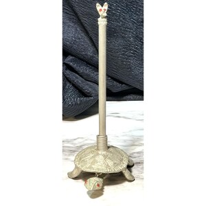 May include: A decorative paper towel holder with a turtle base and a rabbit-shaped top. The holder is made of a light-colored metal and features red accents on the rabbit and turtle. The base is approximately 6 inches in diameter.