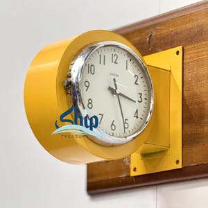 Op de afbeelding: Een vintage Citizen klok met een witte wijzerplaat en zwarte cijfers, geplaatst in een felgele cilindrische behuizing. De klok is gemonteerd op een houten plaat met een gele vierkante lijst. De klok geeft ongeveer 4:15 uur aan.