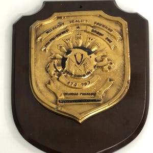 May include: A dark brown wooden shield-shaped plaque featuring a gold-colored metal emblem. The emblem displays the text Military Sealift Command, Prepositioned Group One, Diego Garcia Unit, CTG 79.1, and Invidia Paratus, with a central globe design.