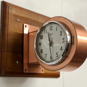 May include: A round clock with a white face and black numerals, set in a copper-colored metal casing. The clock is mounted on a rectangular wooden board. The hands point to approximately 11:00. The clock is a vintage style.