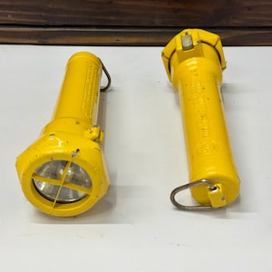 Puede incluir: Dos linternas industriales de color amarillo brillante con cuerpos cilíndricos y cabezales hexagonales. Cada linterna cuenta con una anilla metálica para colgar en la base, una lente de cristal protegida por una rejilla y un interruptor.
