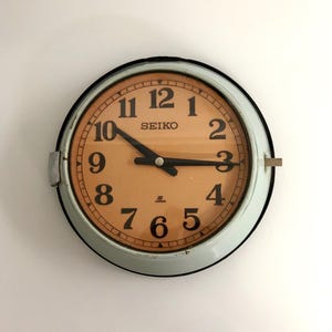 May include: A round, vintage Seiko wall clock with a light blue metal frame and a tan face. Black numerals mark the hours, and black hands indicate the time. The clock face has the Seiko logo and the word "Japan" printed on it.