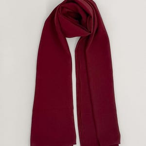 May include: A solid deep burgundy rectangular scarf with a smooth, matte texture. The scarf is draped in a loose, folded loop at the top, with two long, straight ends hanging down vertically, showcasing a consistent dark red color throughout.