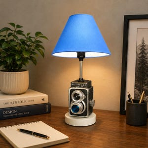 Könnte beinhalten: Eine Tischlampe mit einer vintage schwarzen Ricohflex Model VII zweiäugigen Spiegelreflexkamera als Basis, gekrönt von einem hellblauen, konischen Stofflampenschirm. Das Kameragehäuse ist metallisch schwarz mit silbernen Akzenten auf einem weißen Sockel.