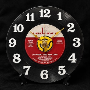 May include: A wall clock crafted from a black vinyl record featuring white numerals from 1 to 12. The center label is red and white with the text Cadence, Andy Williams, and It Doesn't Take Very Long. It has yellow clock hands and a yellow center adapter.