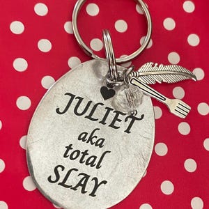 Puede incluir: Un llavero plateado con la inscripción "JULIET aka total SLAY" grabada en un colgante ovalado. El llavero incluye un dije de pluma, un dije de tenedor y un pequeño corazón. El fondo es rojo con lunares blancos.