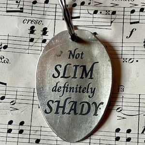 Puede incluir: Un llavero ovalado plateado con la inscripción "Not SLIM definitely SHADY" en negro. El llavero está unido a un anillo plateado. El fondo es una partitura musical antigua.