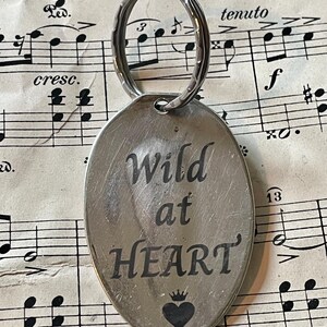 Puede incluir: Llavero ovalado plateado con las palabras "Wild at HEART" grabadas en negro. Un pequeño corazón negro con una corona está debajo del texto. El llavero tiene un anillo plateado en la parte superior para sujetar las llaves.