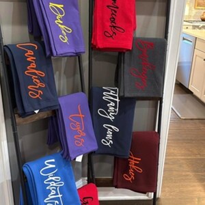 Pode incluir: Exibição de vários cobertores de lã polar em diferentes cores, cada um bordado com o nome de uma equipa em fonte cursiva. Os cobertores estão dispostos numa escada decorativa. As cores incluem roxo, vermelho, azul marinho, cinzento, azul e bordô.