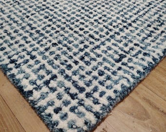 May include: A close-up shot of a patterned rug. The rug has a geometric design in shades of white and blue. The corner of the rug is visible, showing its thick, textured pile. The rug is placed on a wooden floor.